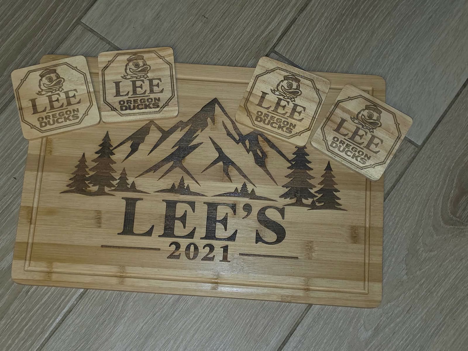 Walnut-toned bamboo cutting board engraved with mountains, pines, and LEE'S 2021, with four matching engraved coasters